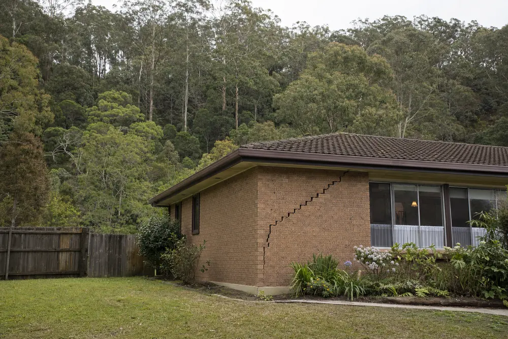 A brick house with a long stair-step crack down its corner wall, in front of a steep tree-covered hill — underpinning, Wollongong. Illustrative example.