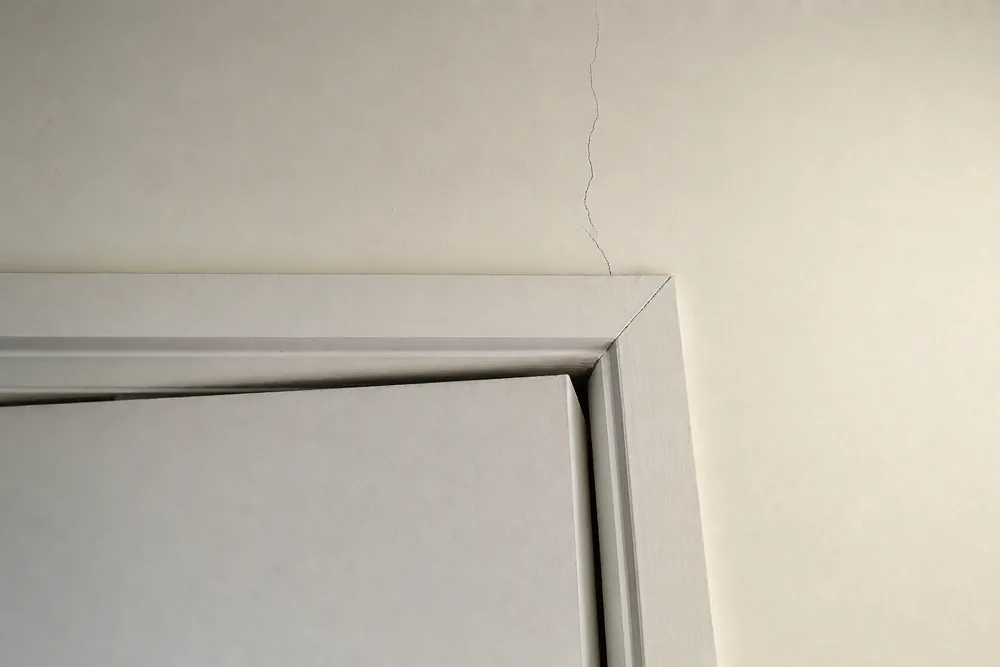 The top corner of an interior door frame with a wedge-shaped gap beside the door and a hairline crack in the wall above — foundation repair, Wollongong. Illustrative example.