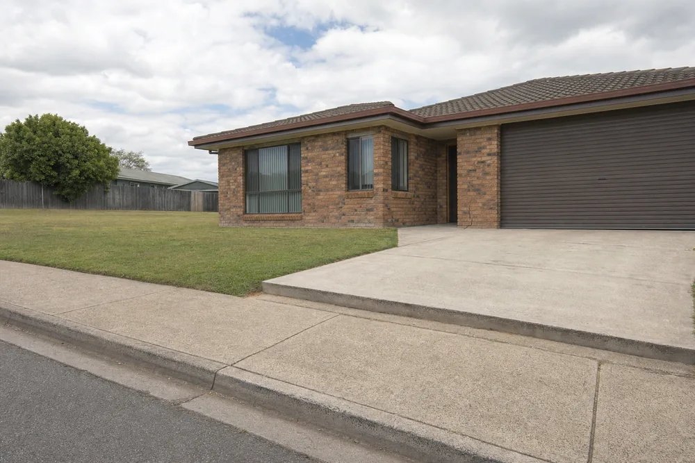 A low brick house with a garage on a flat lawn, a concrete driveway with a raised step at its edge, and a thin crack in the wall beside the garage door — underpinning, Wollongong. Illustrative example.